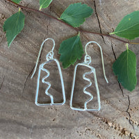 Pair of open work silver earrings with a squiggle down the center on a wooden surface with leaves
