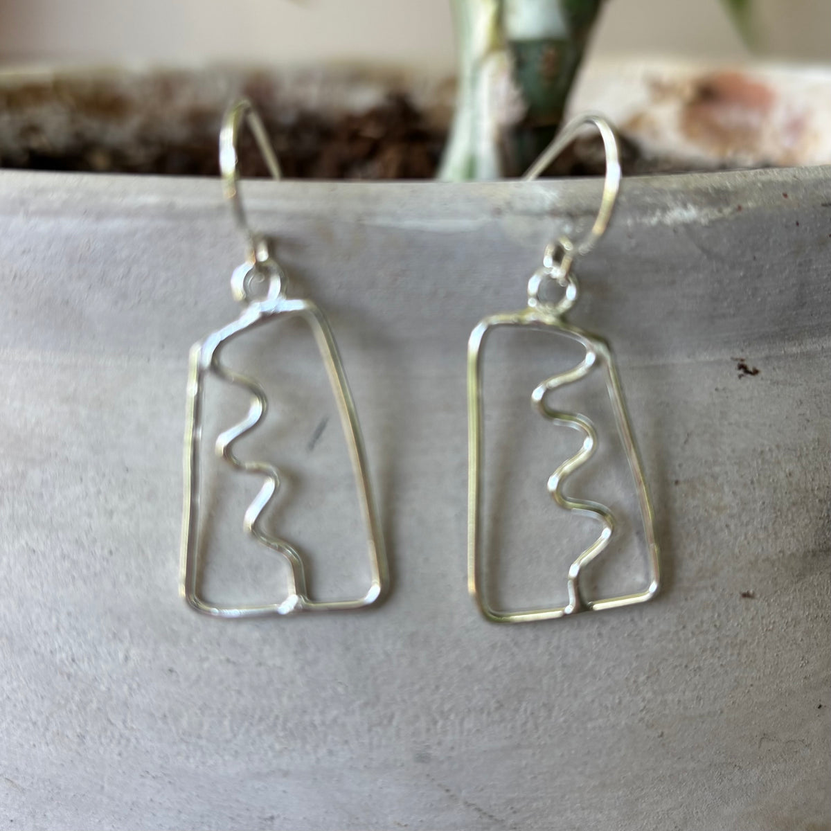 Silver abstract earrings with a squiggle down the center on a textured surface with a plant in the background