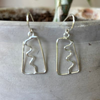 Silver abstract earrings with a squiggle down the center on a textured surface with a plant in the background