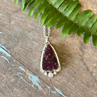 Sterling silver necklace with a purple druzy pendant on a wooden surface with a green leaf.