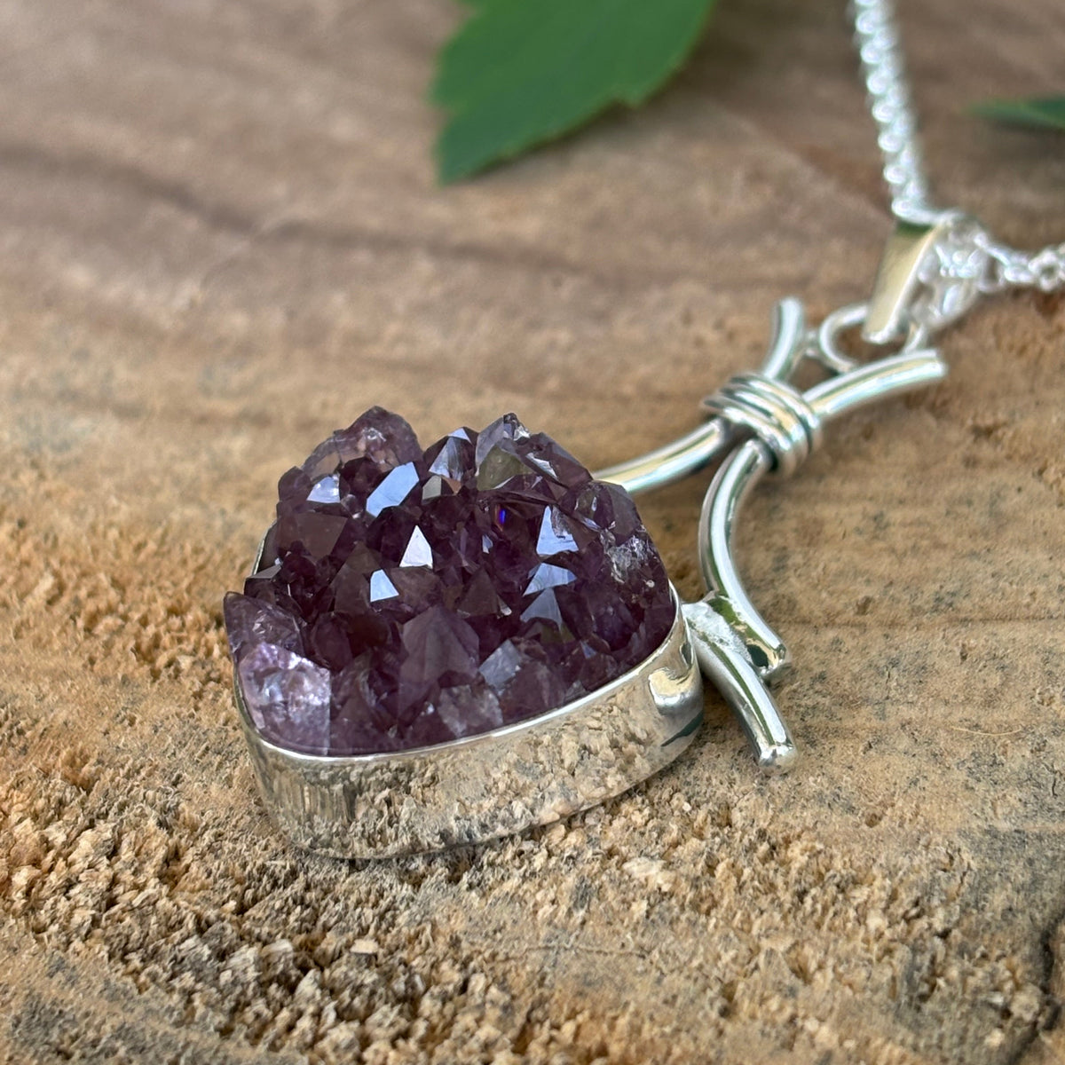 side view of Druzy Amethyst pendant on a silver chain with a green leaf on a wooden surface
