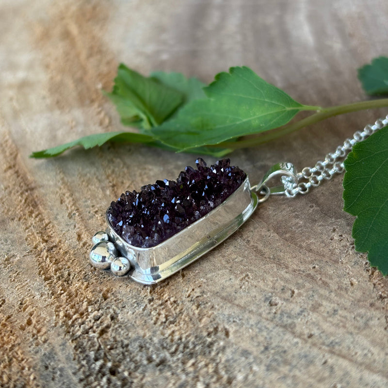 Sterling silver necklace with a purple druzy pendant on a wooden surface with a green leaf.