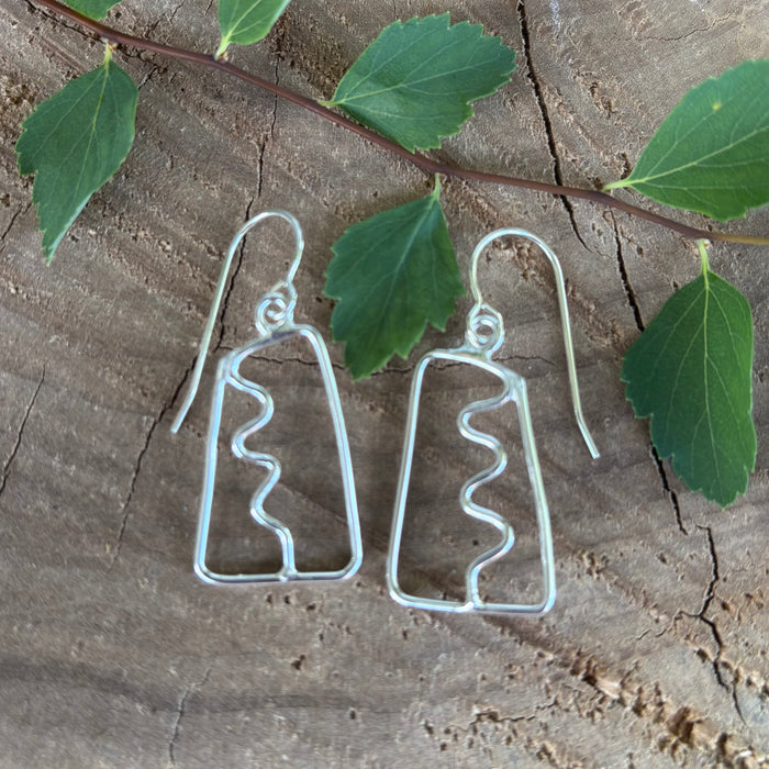Pair of open work silver earrings with a squiggle down the center on a wooden surface with leaves