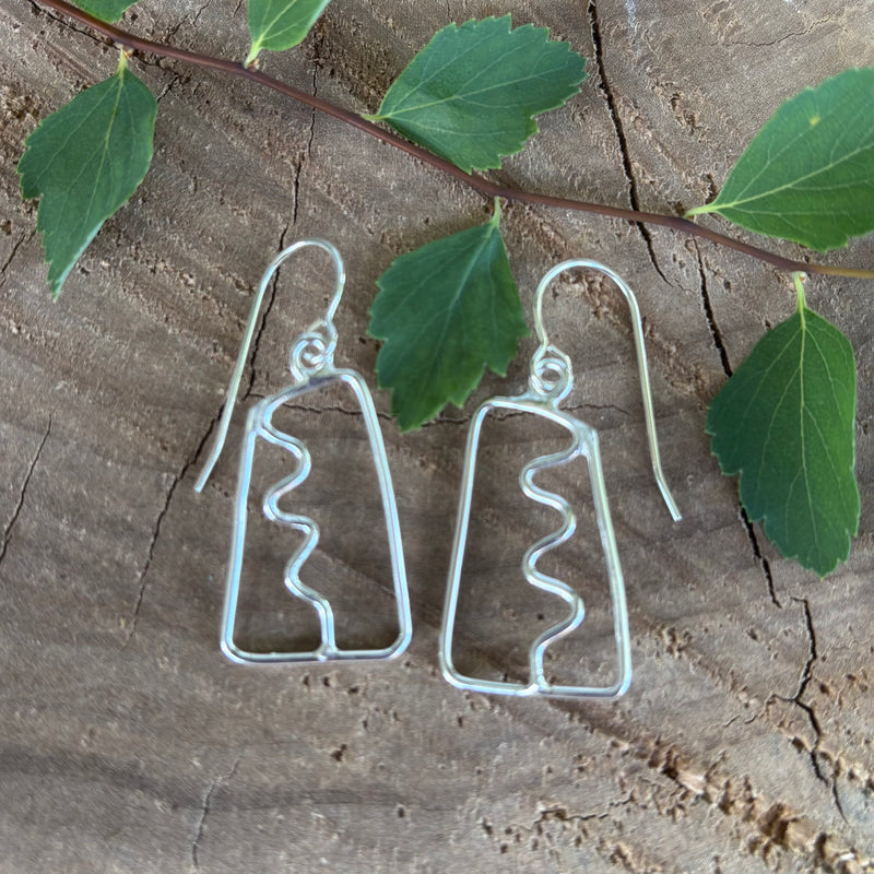 Pair of open work silver earrings with a squiggle down the center on a wooden surface with leaves