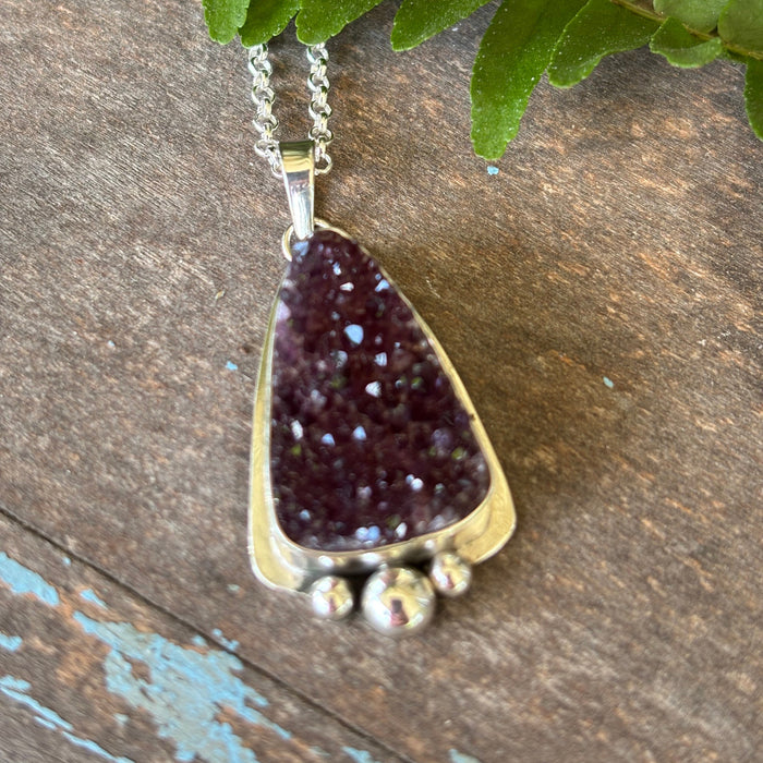 Sterling silver necklace with a purple druzy pendant on a wooden surface with a green leaf.