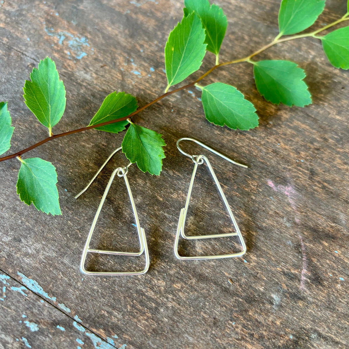 Silver triangle earrings on a wooden surface with green leaves