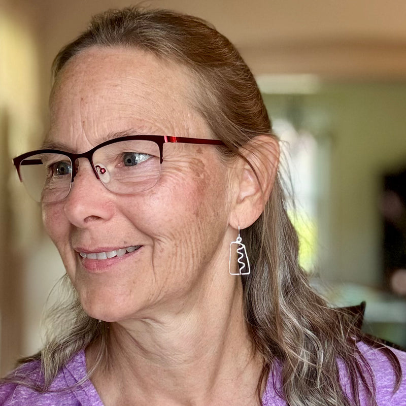 Woman wearing abstract silver earrings with a squiggle down the center. She is wearing glasses and a purple shirt.