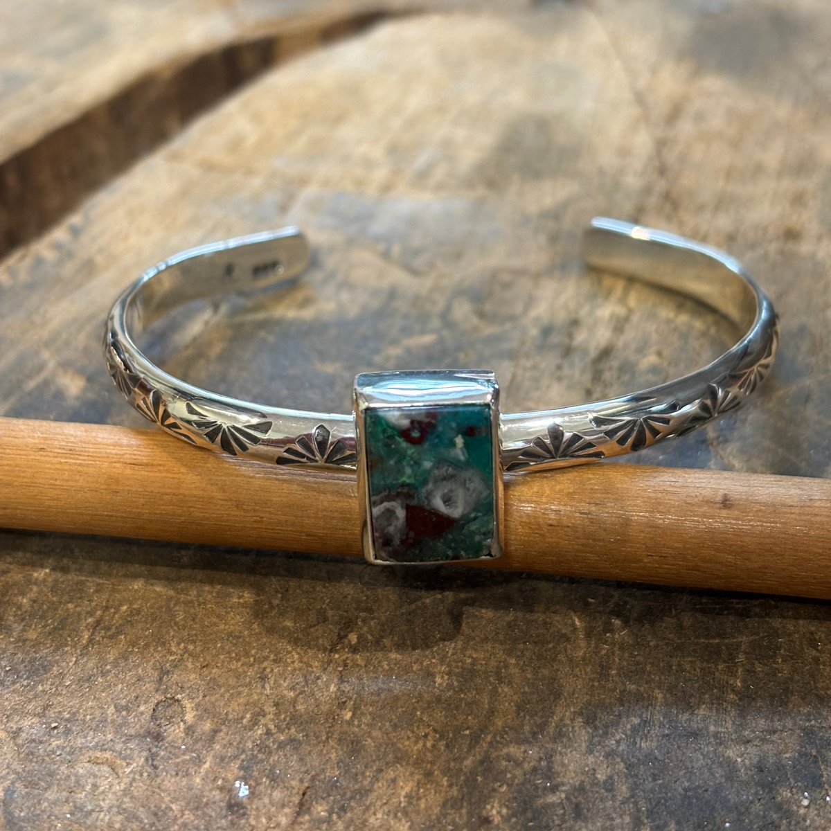 A sterling silver cuff bracelet with engraved floral details and a rectangular turquoise stone centerpiece, resting on a wooden workbench.